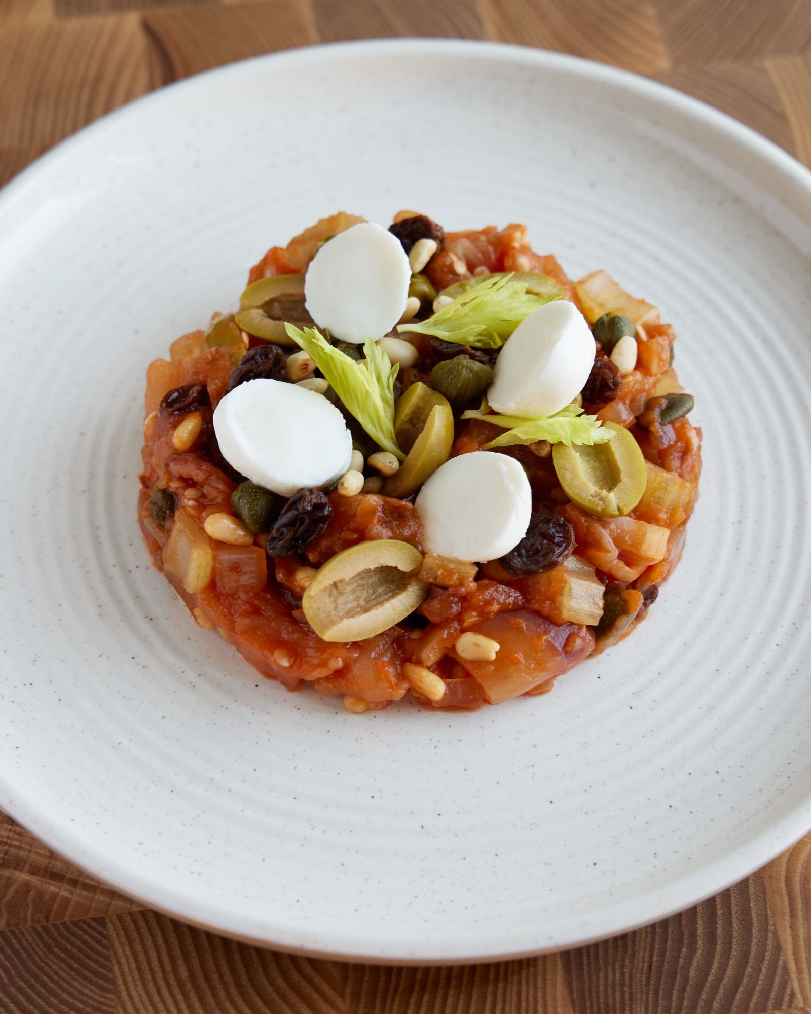 Caponata siciliana di melanzane con olive, capperi, pinoli e uvetta, servita in un piatto bianco.