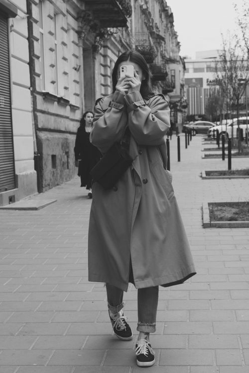 Free Monochrome image of a woman in a trench coat taking a photo with a cellphone on a city sidewalk. Stock Photo
