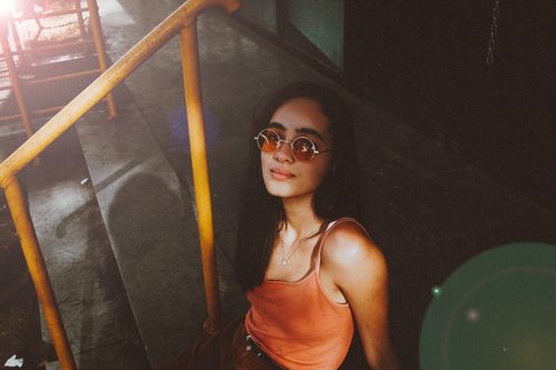 Free A fashionable woman in an orange tank top and sunglasses sitting on stairs, creating a warm and moody atmosphere. Stock Photo