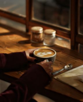 A person holds a cappuccino with latte art near a window in a cozy coffee shop.