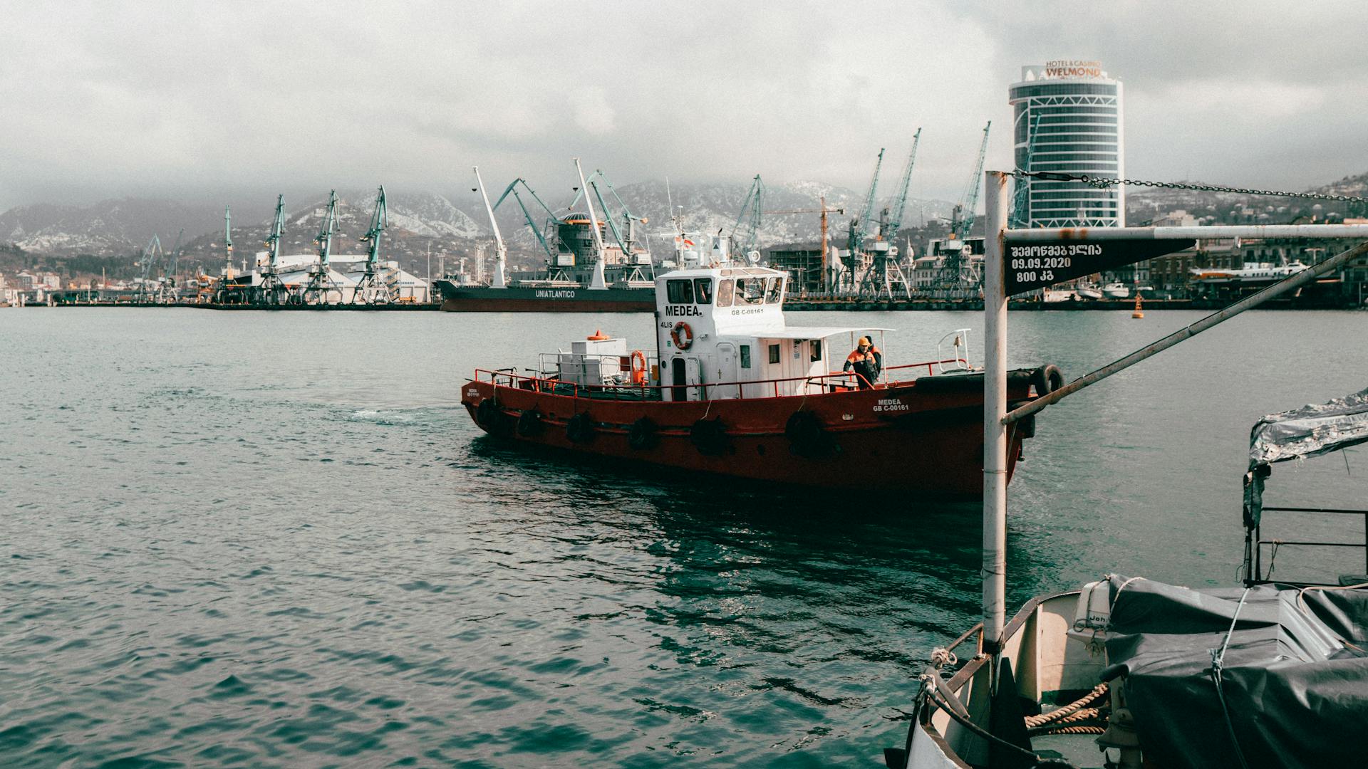 A tugboat navigating the bustling Batumi port, featuring cranes and industrial structures.