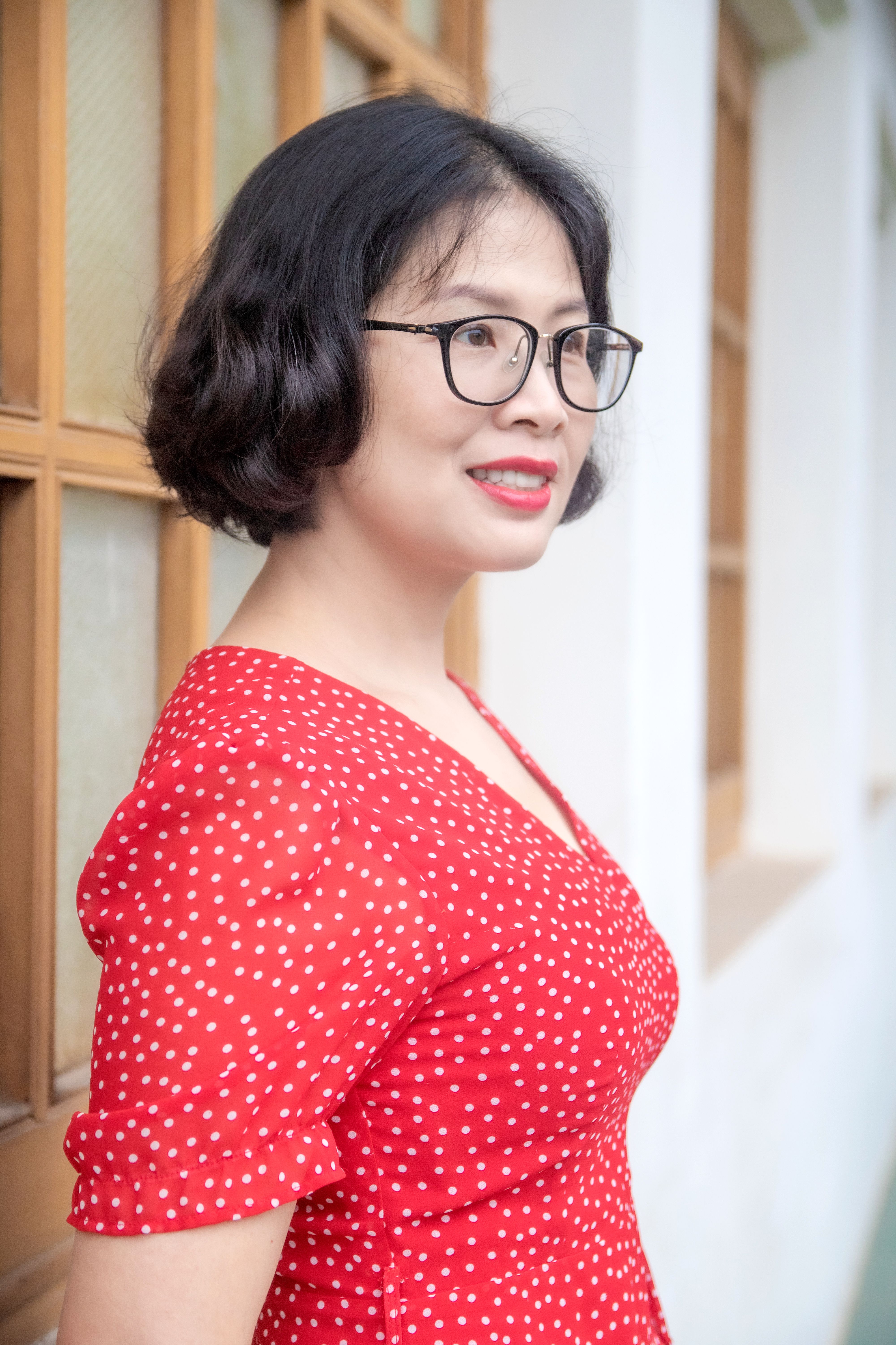 A cheerful woman in a red polka dot dress and eyeglasses standing by a window.
