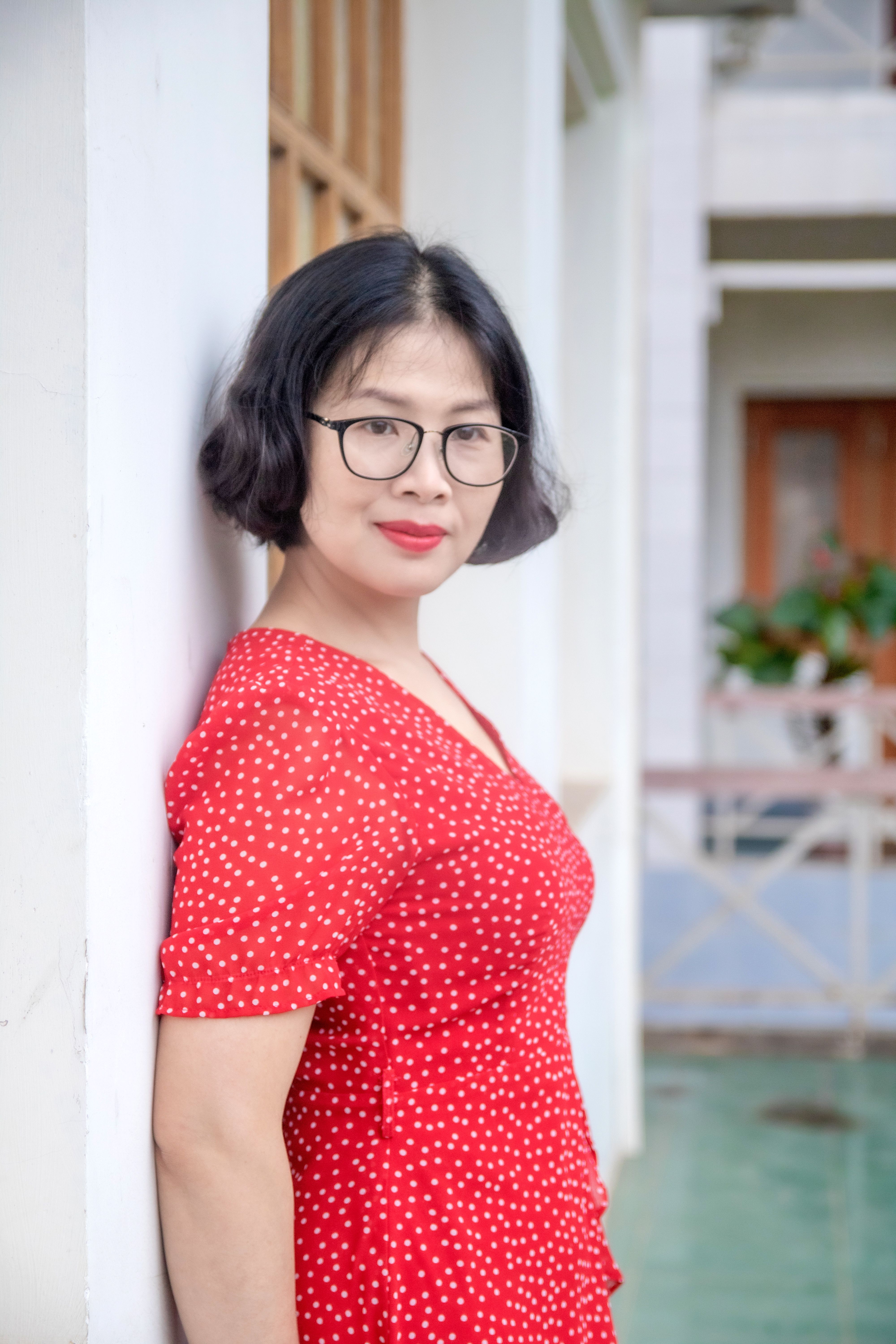 Portrait of a woman leaning against a wall, wearing eyeglasses and a red polka dot dress, smiling confidently.