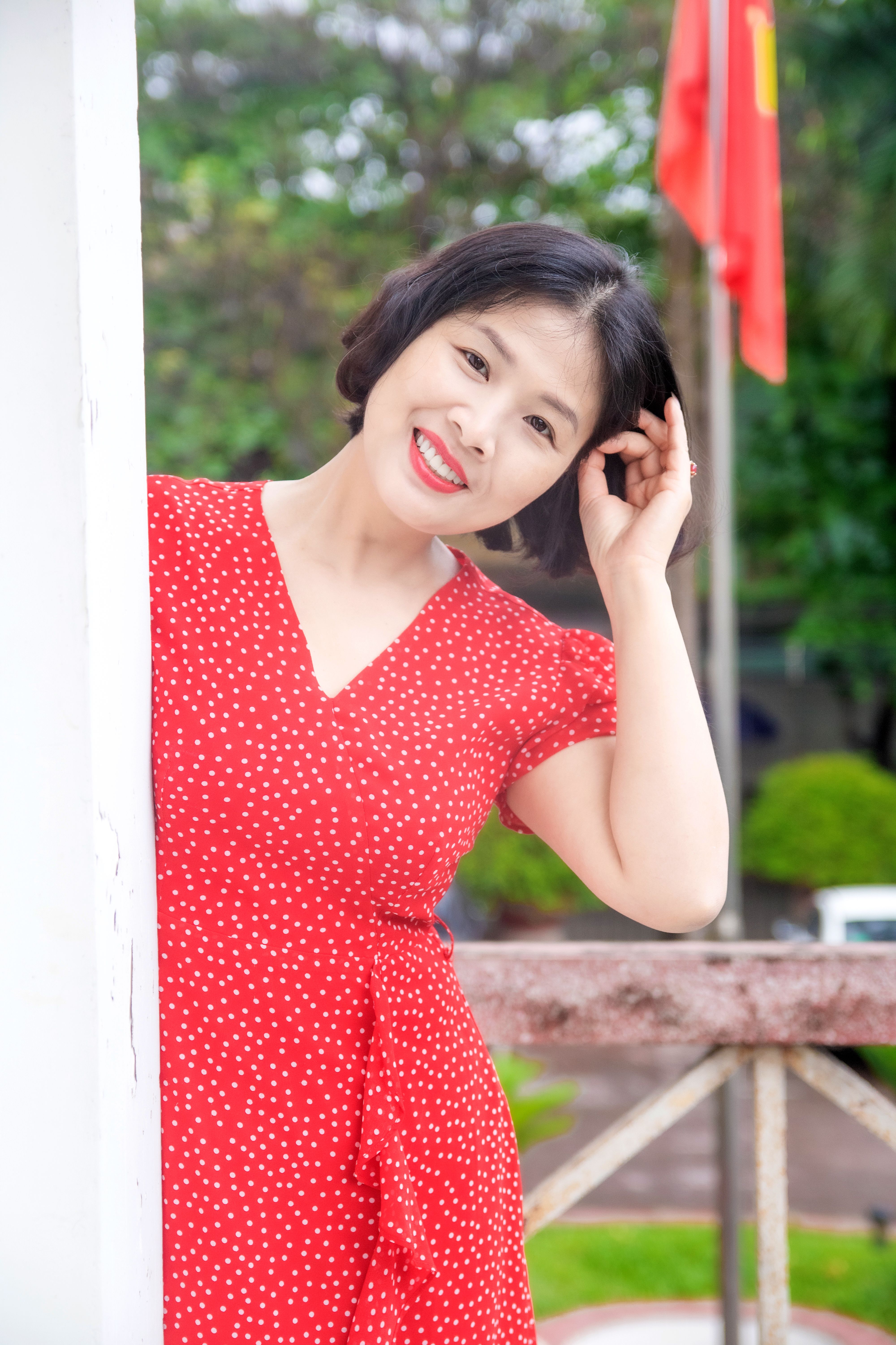 Cheerful woman in a red polka dot dress smiling outdoors in a lush green setting.