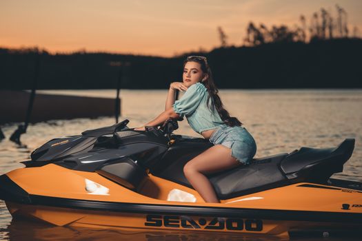 A fashionable young woman poses on a jet ski, surrounded by a scenic sunset over calm waters, exuding elegance.
