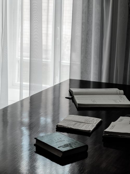 Free A peaceful reading corner with books on a wooden table by a window with sheer curtains. Stock Photo