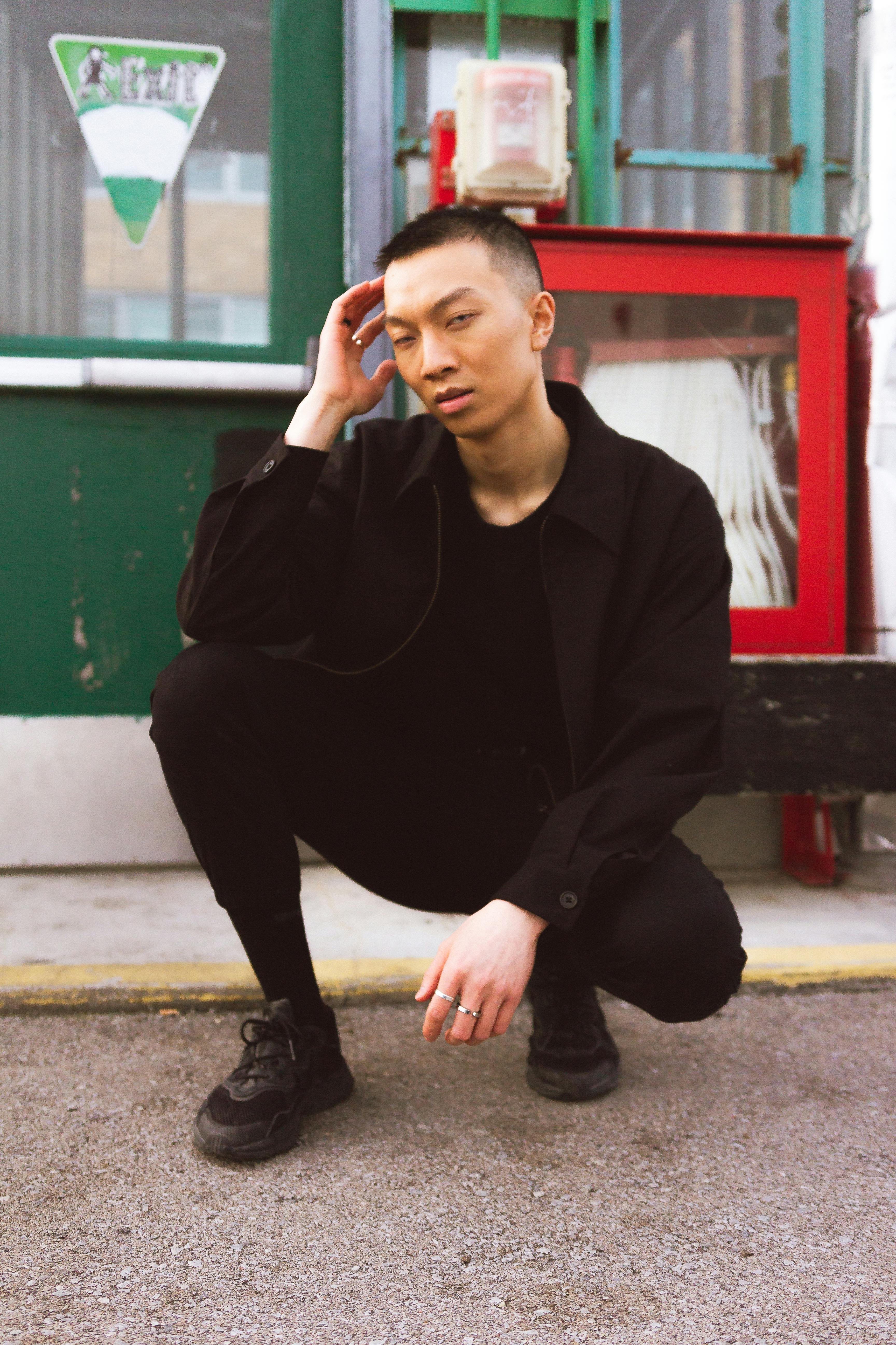 A young man in black poses thoughtfully outdoors, conveying a trendy and introspective mood.