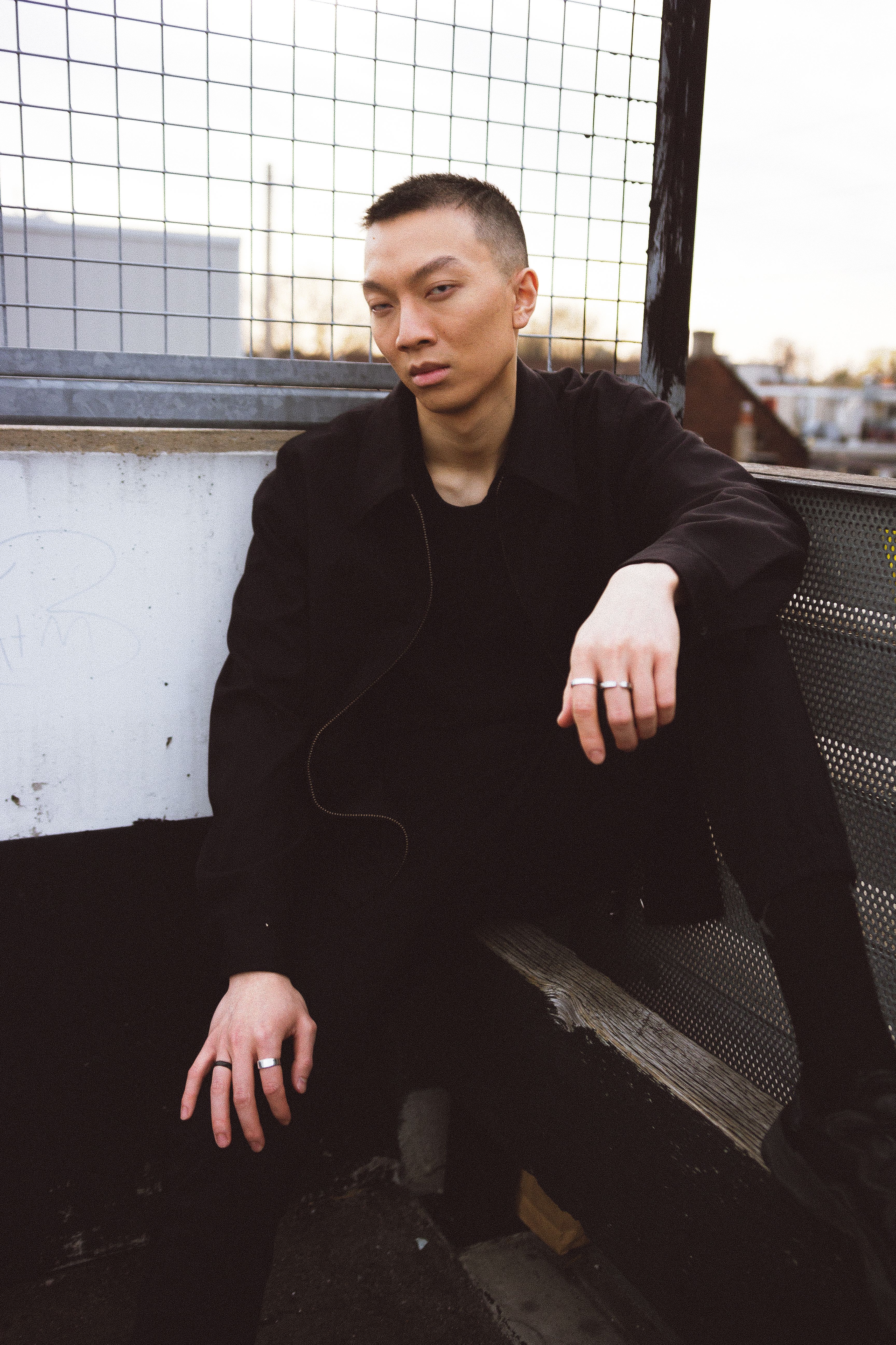 A fashionable man in black attire poses seriously on a rooftop, exuding a modern urban vibe.