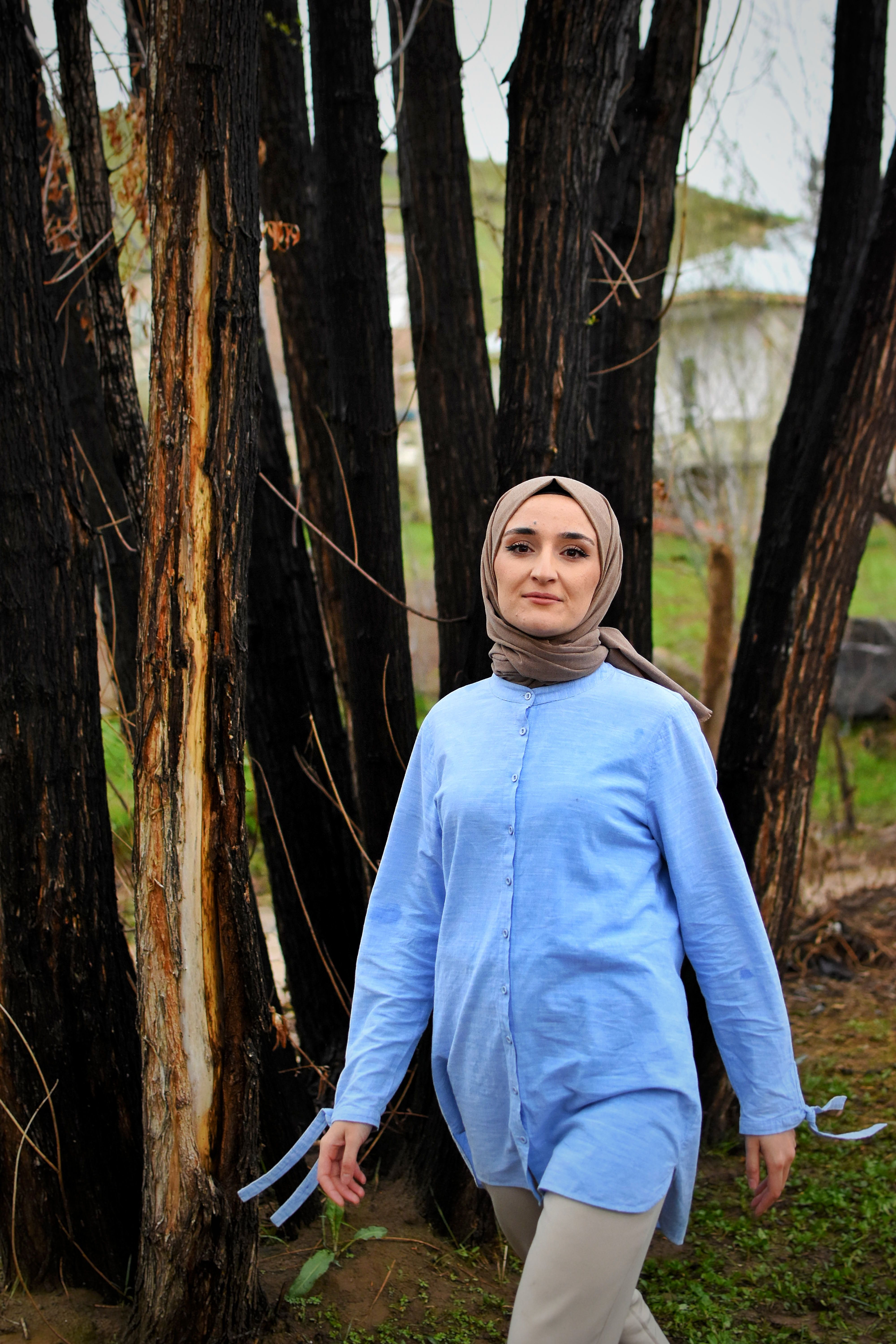 A woman wearing a blue outfit and hijab walks outdoors near trees.