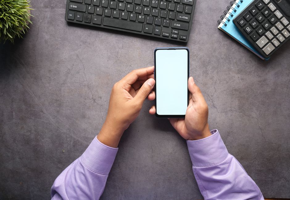 Man in purple shirt holding a smartphone with blank screen at office desk with keyboard, notebook, and calculator.