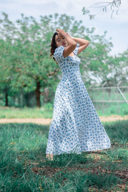 Free Beautiful Asian woman posing elegantly in a floral dress amidst lush green park during summer. Stock Photo