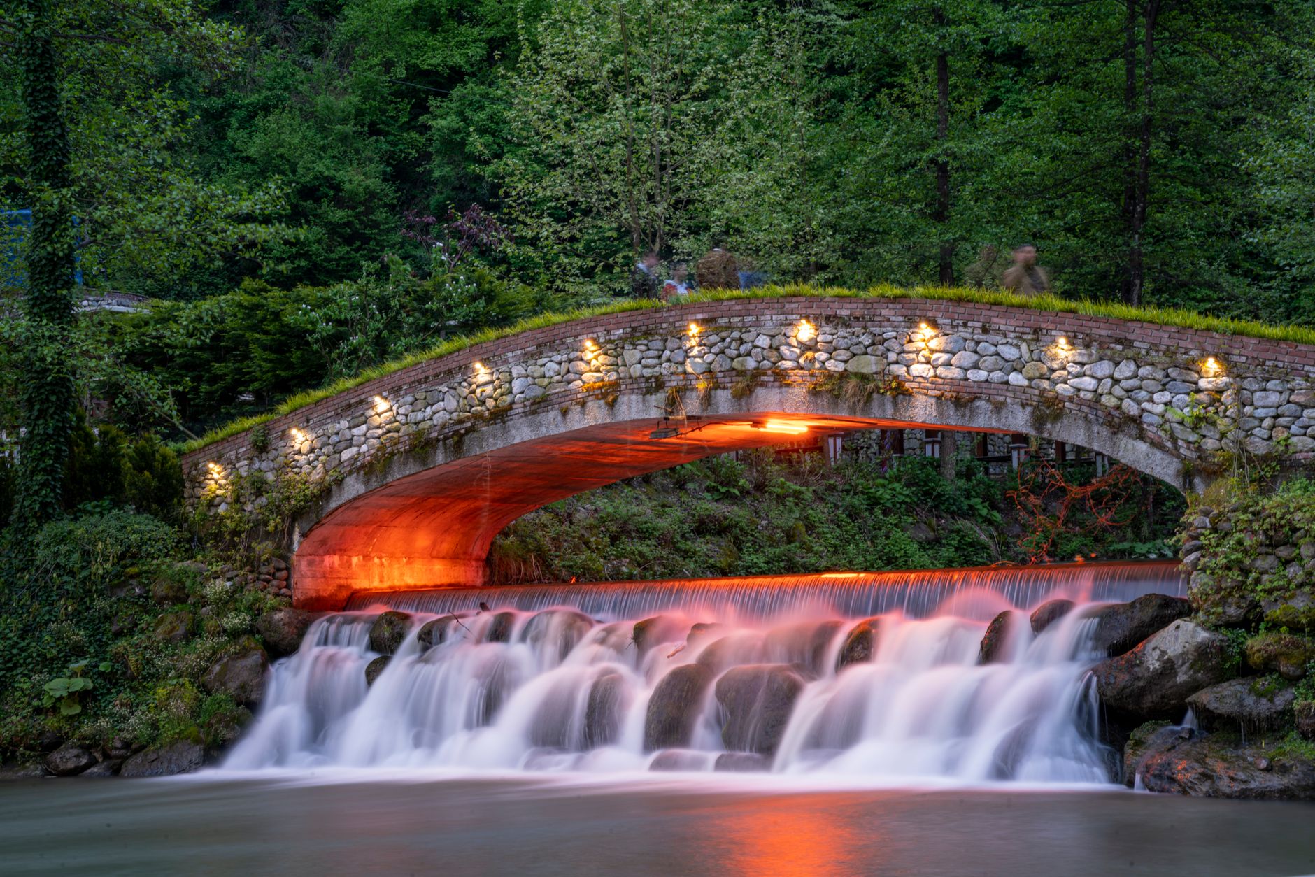 A scenic illuminated stone bridge over a waterfall in a lush park in Sakarya, Turkey, creating a tranquil nature scene.
