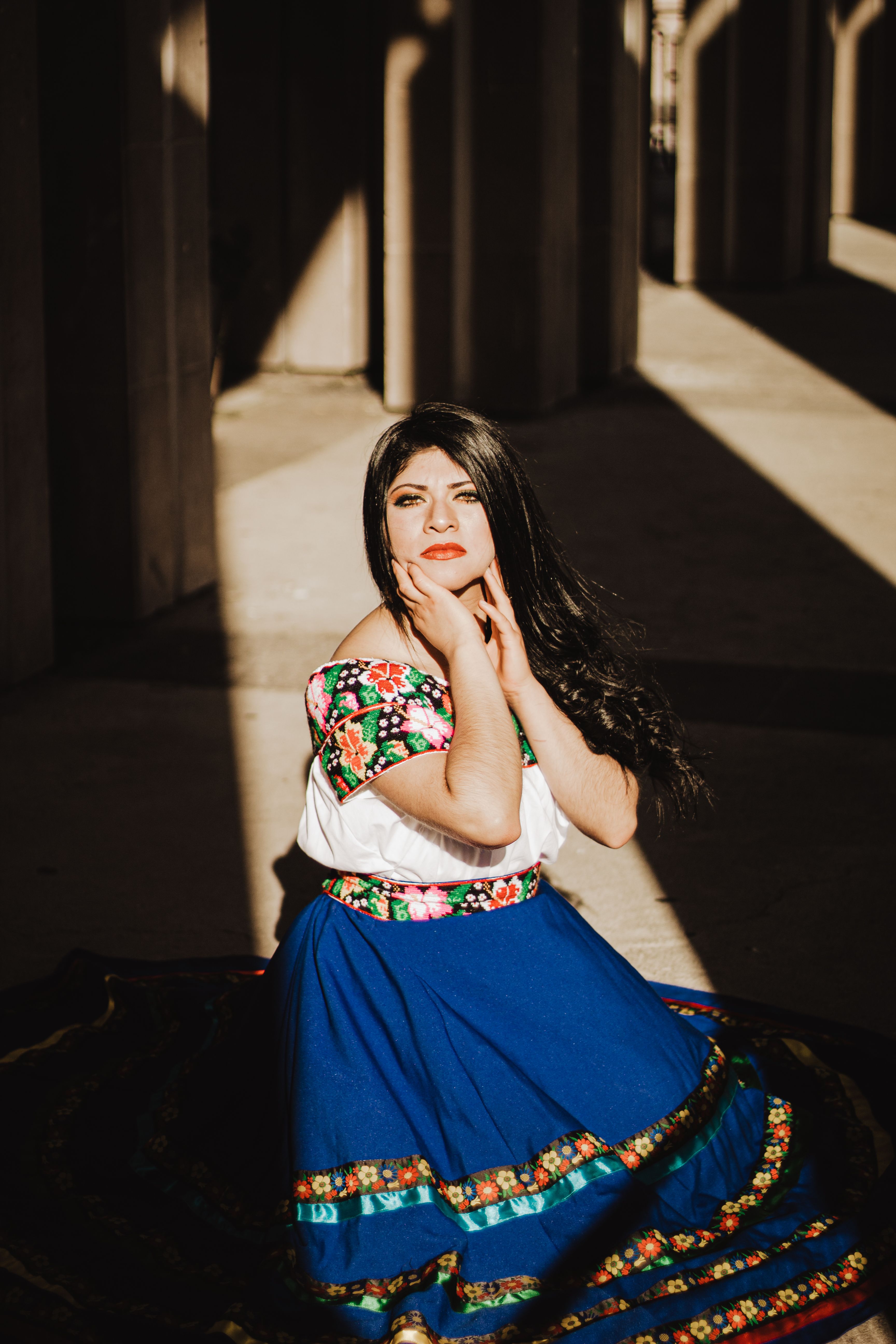 Portrait of a woman in colorful traditional Mexican attire posing thoughtfully with hands on chin.