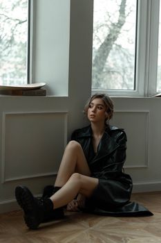 Elegant woman in black leather jacket sitting gracefully on a wooden floor by a window.