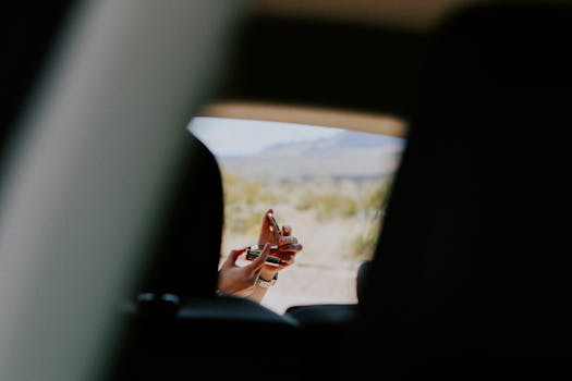 A woman applying red lipstick using a compact mirror outside a vehicle in a desert setting.