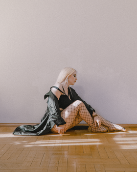 Stylish blonde woman in fishnet tights poses gracefully indoors against a minimalist backdrop, Saint Petersburg.