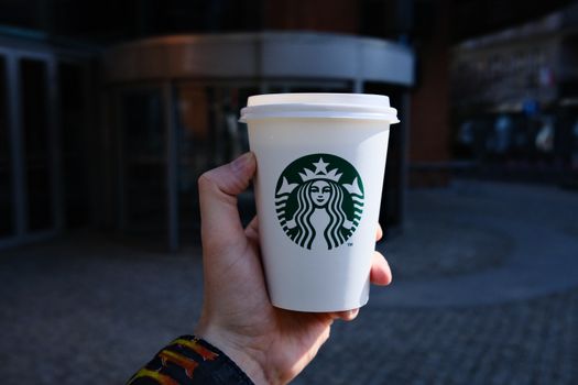 A hand holds a disposable coffee cup with a logo outdoors in Poznań, Poland.