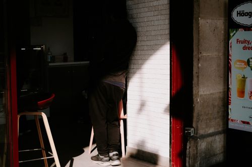 Free A shadowy figure stands beside a storefront displaying advertisements in soft daylight. Stock Photo