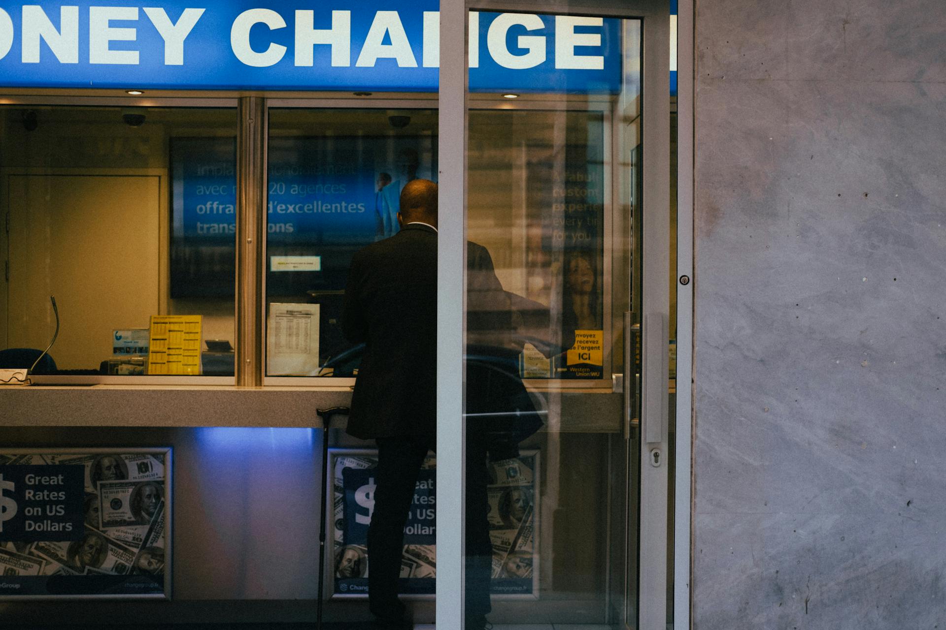 Man at a currency exchange office window, showing currency rates inside a bustling city.