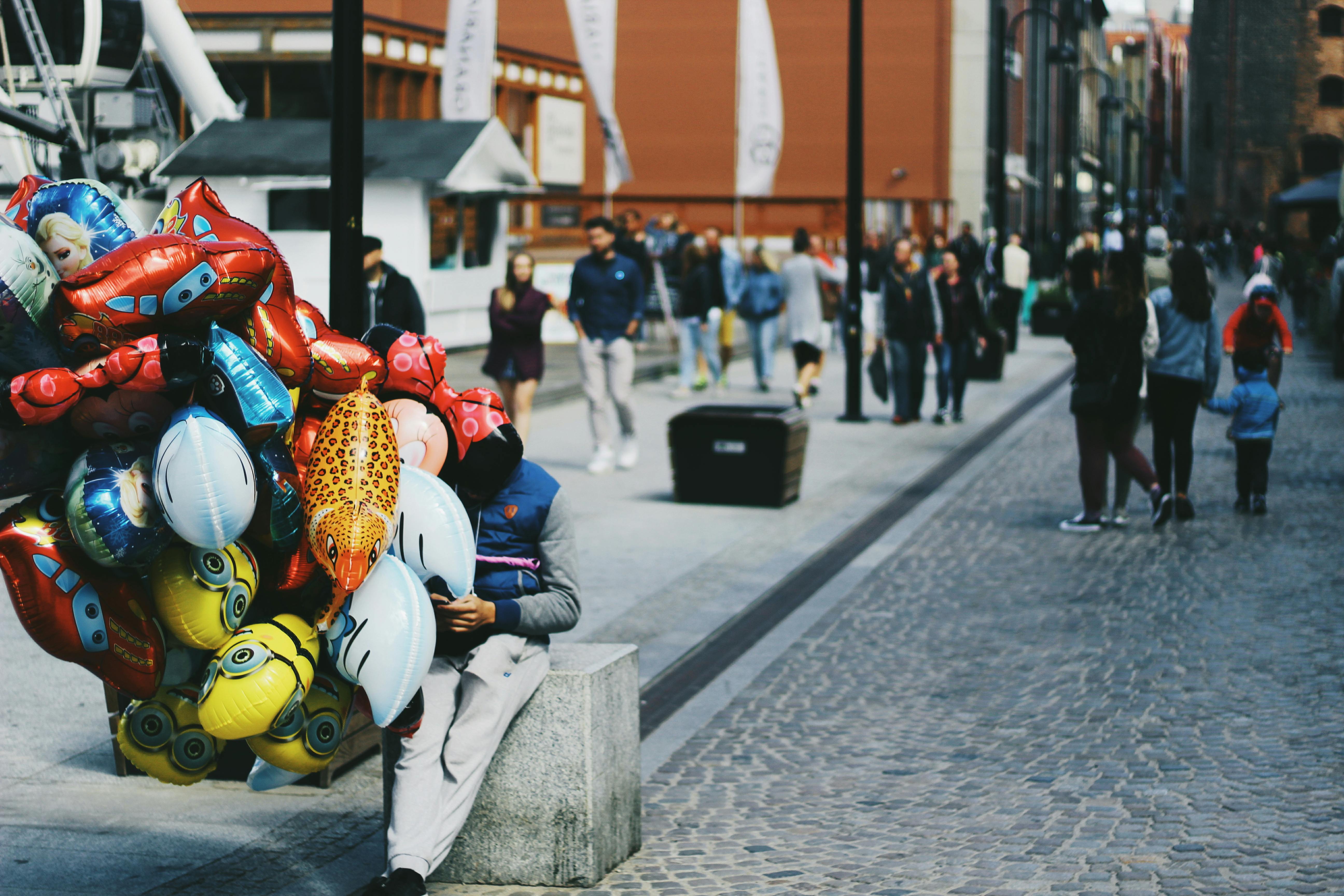 热闹的户外市场街道上，五颜六色的气球摆放着，行人熙熙攘攘