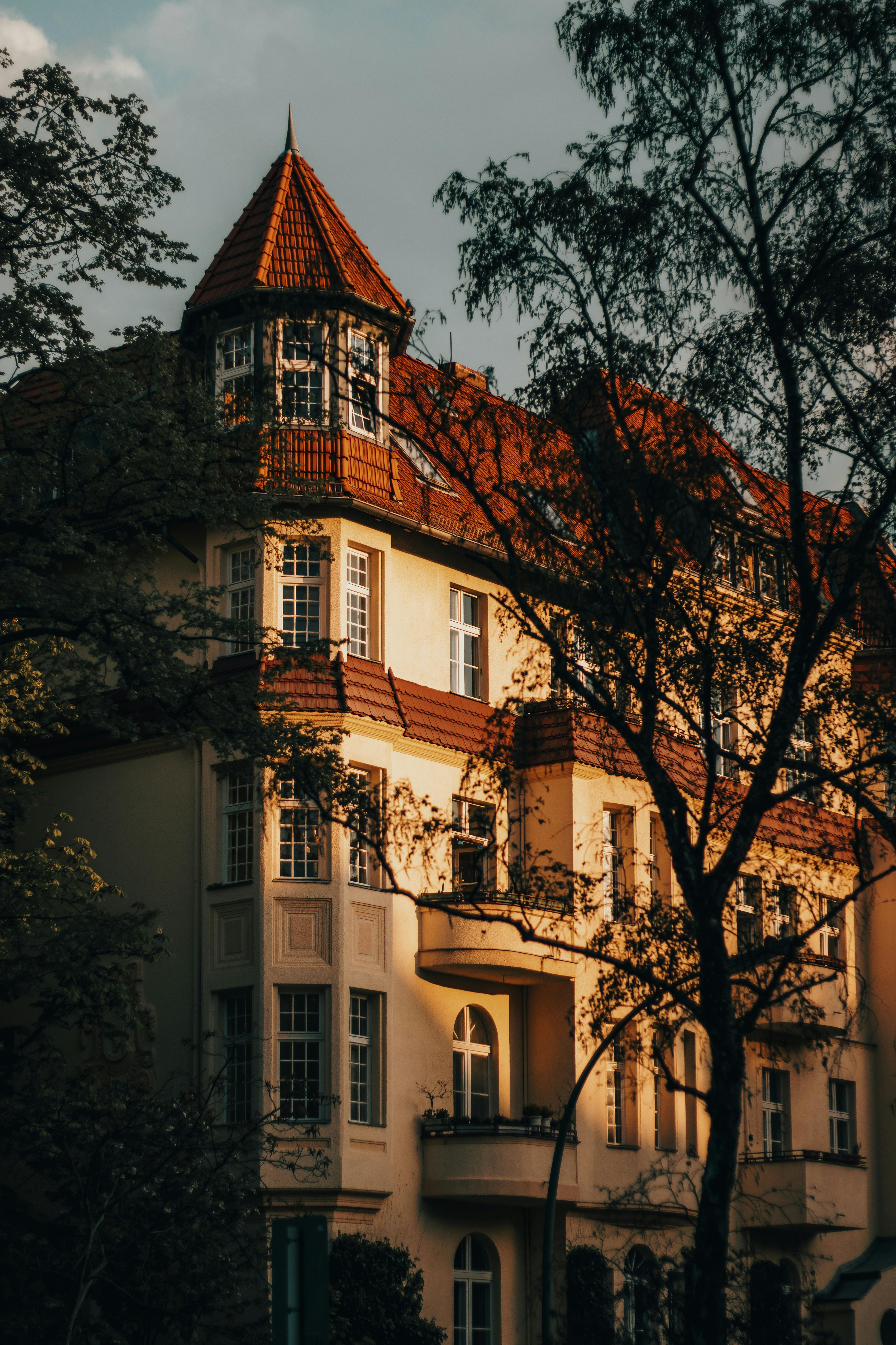 Sunlit European residential building with classic facade and red roof surrounded by trees.