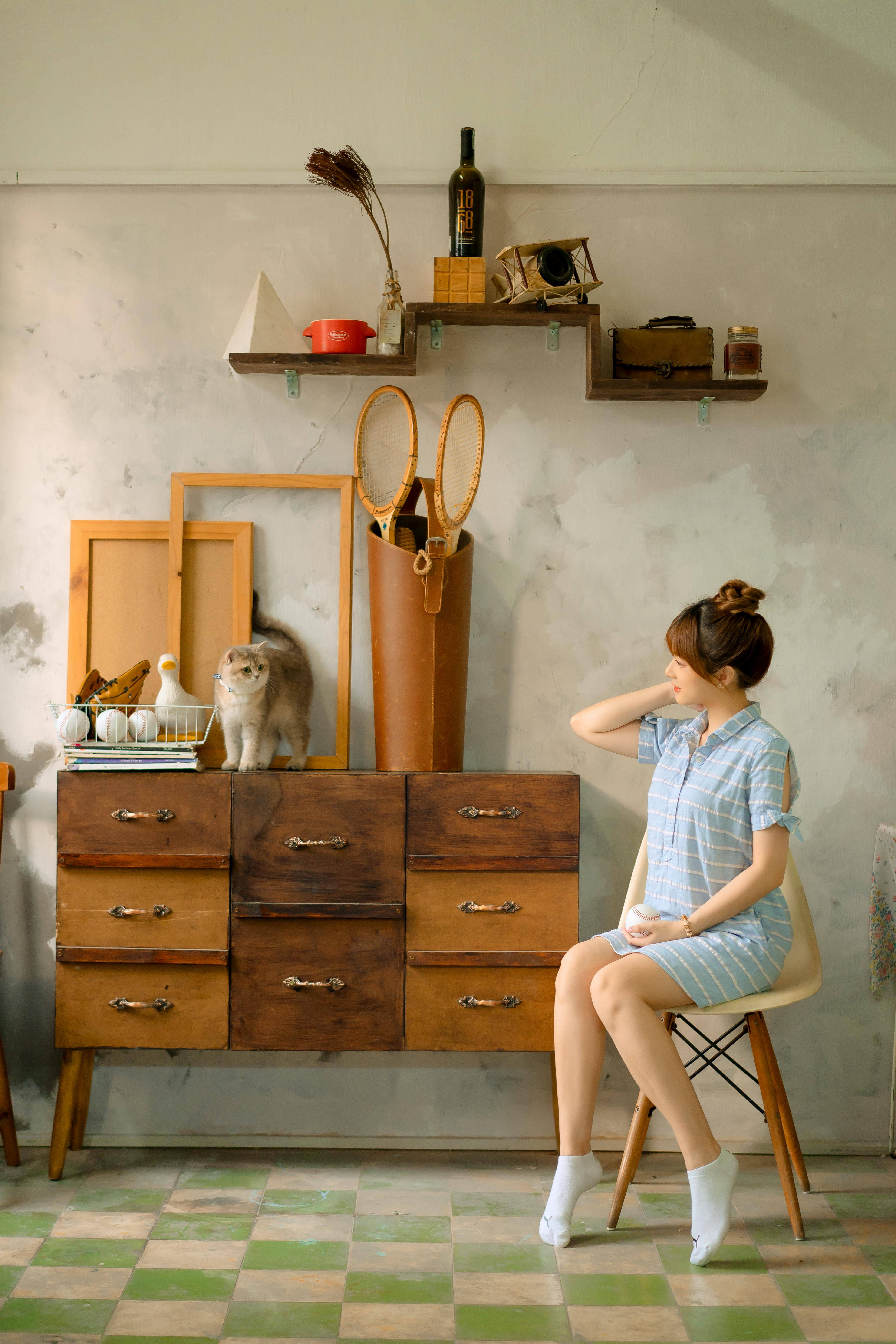 woman sitting on a chair and looking at a cat walking on a cabinet