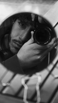 Black and white portrait of a man reflected in a round mirror holding a camera.