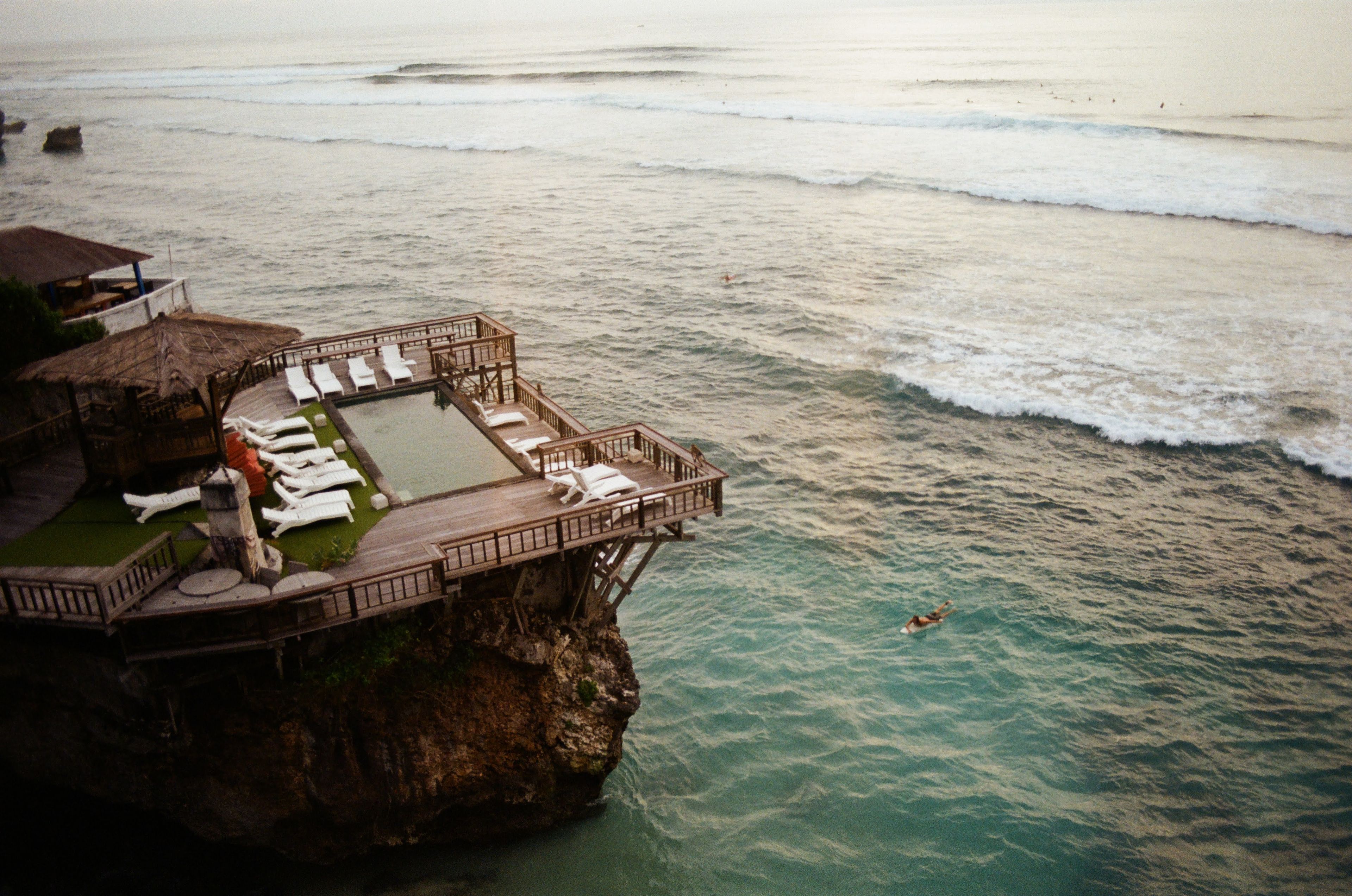 Aerial view of a luxurious cliffside resort in Bali, Indonesia, with ocean and swimmers below.