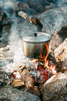 Close-up of a metal pot simmering over an open campfire with smoke and flames.