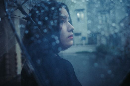 Artistic portrait of a woman holding an umbrella on a rainy day, with water drops and a moody atmosphere.