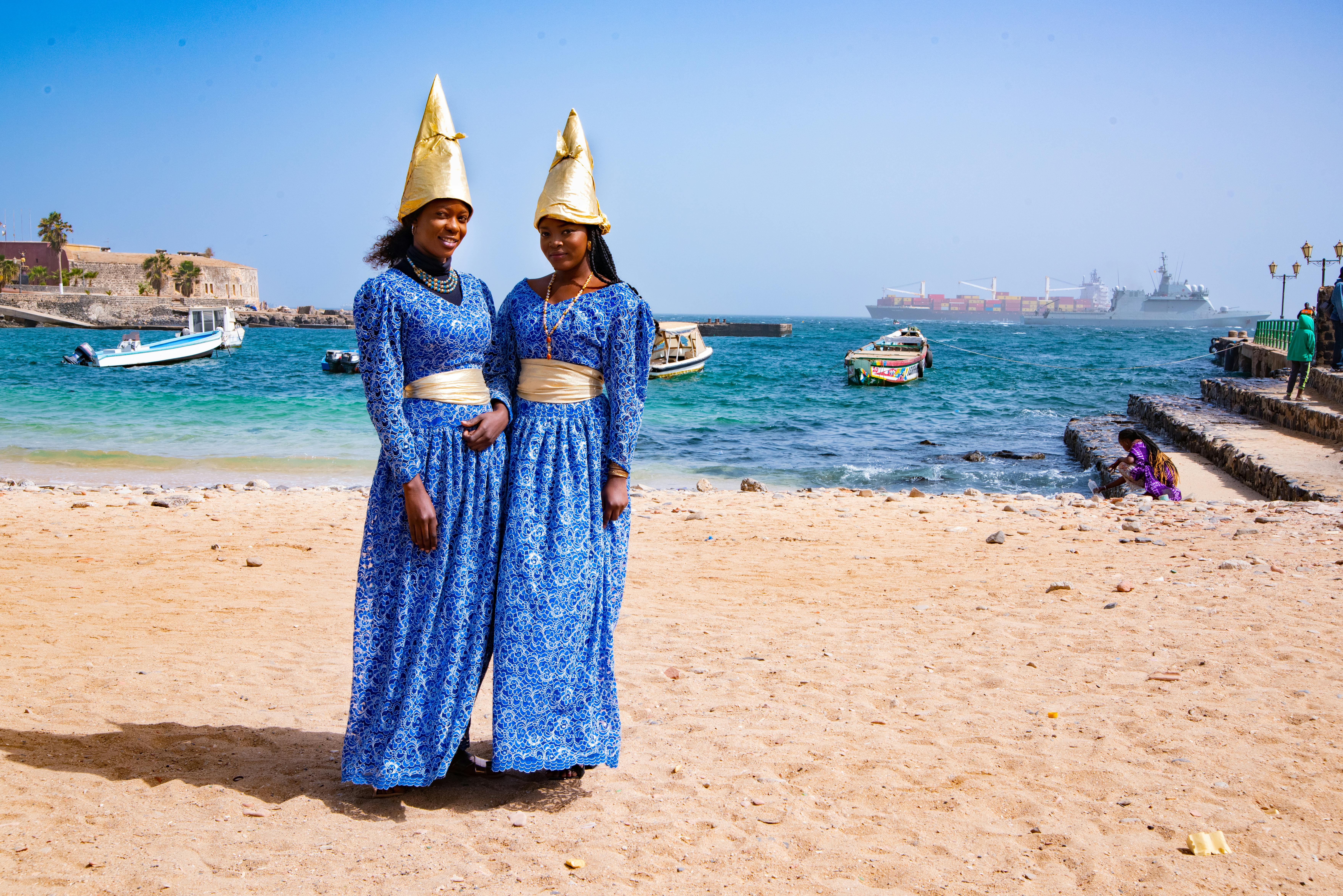 Two women in blue traditional dresses with cone hats on a sunny beach in Dakar, Senegal.