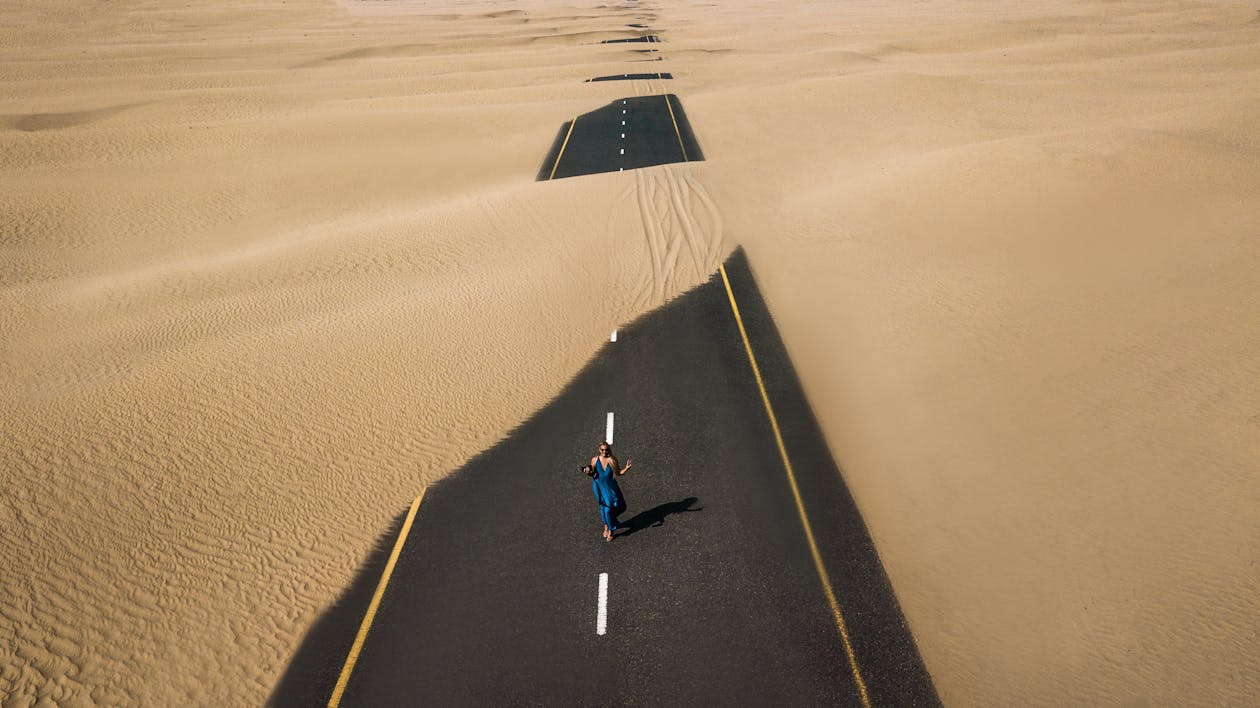 Free A lone woman walking on a sand-covered road in Dubai's desert, captured from above. Stock Photo