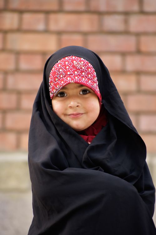 Free Cute little girl wearing a hijab with a bright smile, standing against a brick wall background. Stock Photo