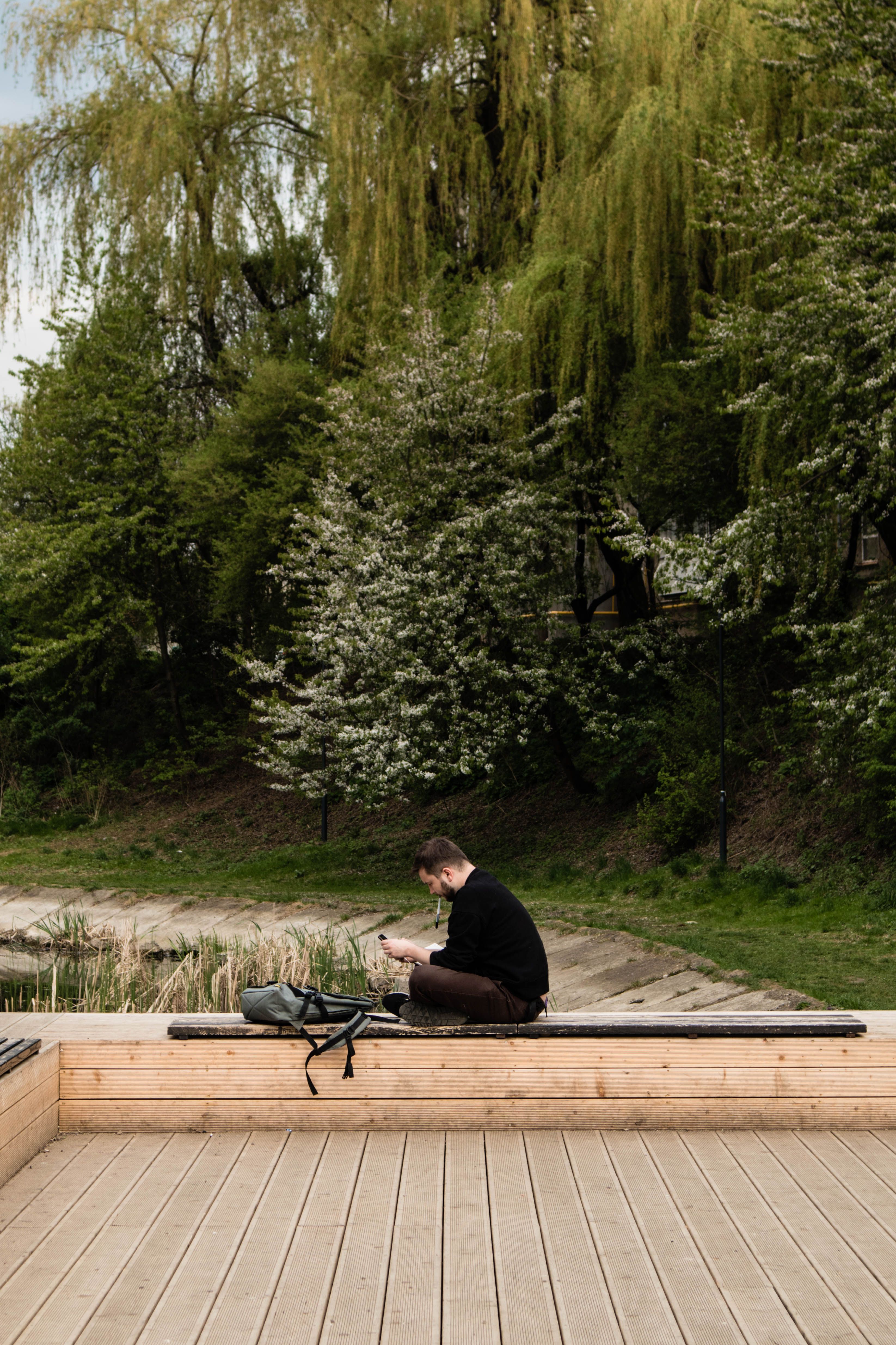 A man in a park using a smartphone while sitting on a bench beside a pond.