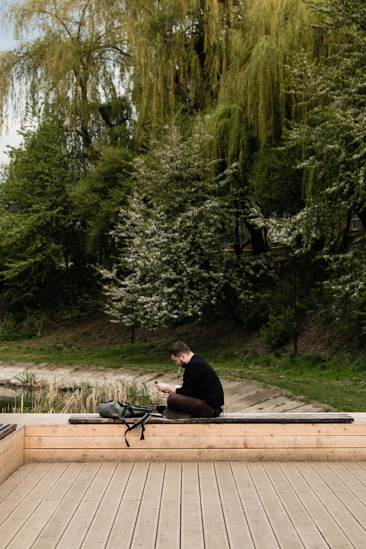 Man In Black Sweater Sitting On A Bench By A Pond In A Park