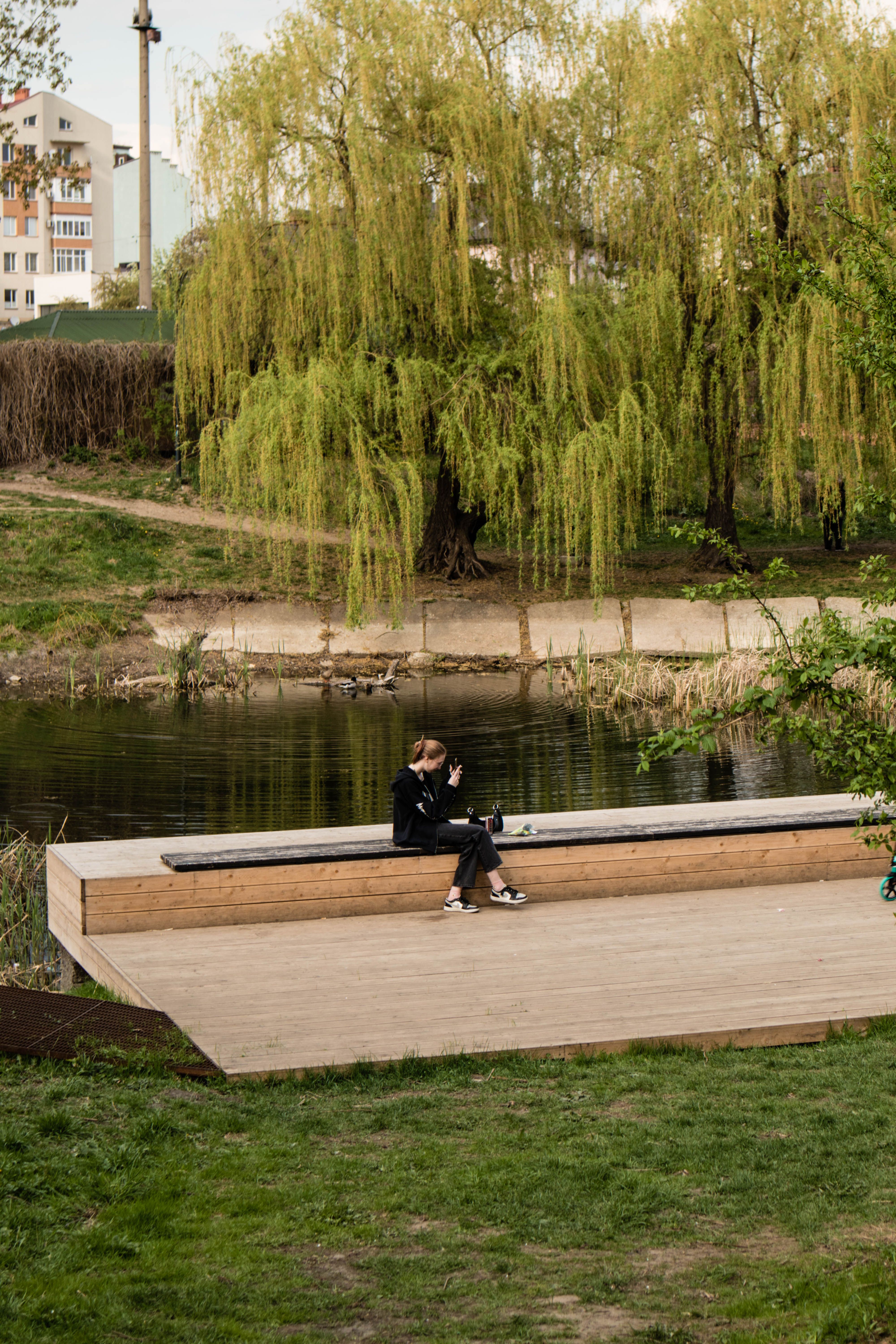 A woman enjoys leisure time on a bench in a serene park beside a tranquil river, surrounded by lush greenery.