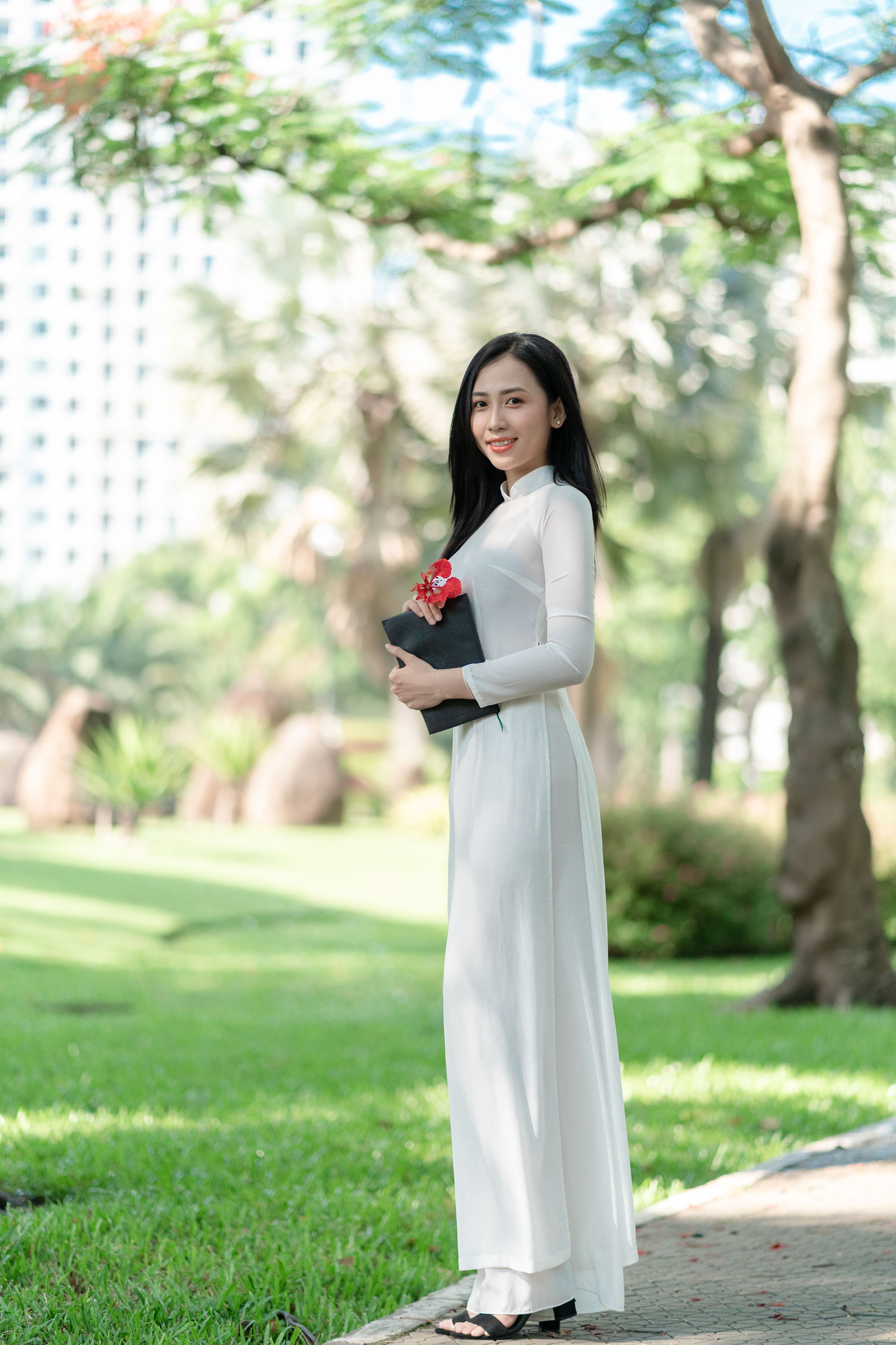 Elegant Asian woman in white ao dai dress stands gracefully outdoors in a scenic park setting.