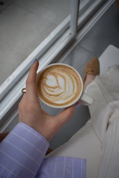 A close-up of a hand holding a latte with intricate latte art in a cozy setting.