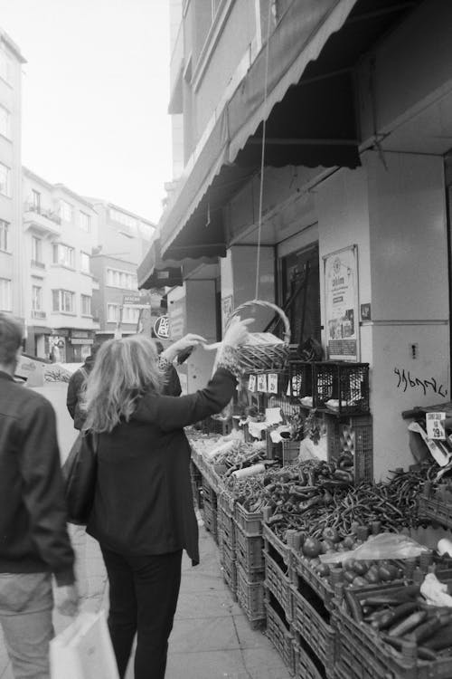 Free Grayscale Photo of Woman in Black Coat Standing in Front of Vegetable Stand Stock Photo