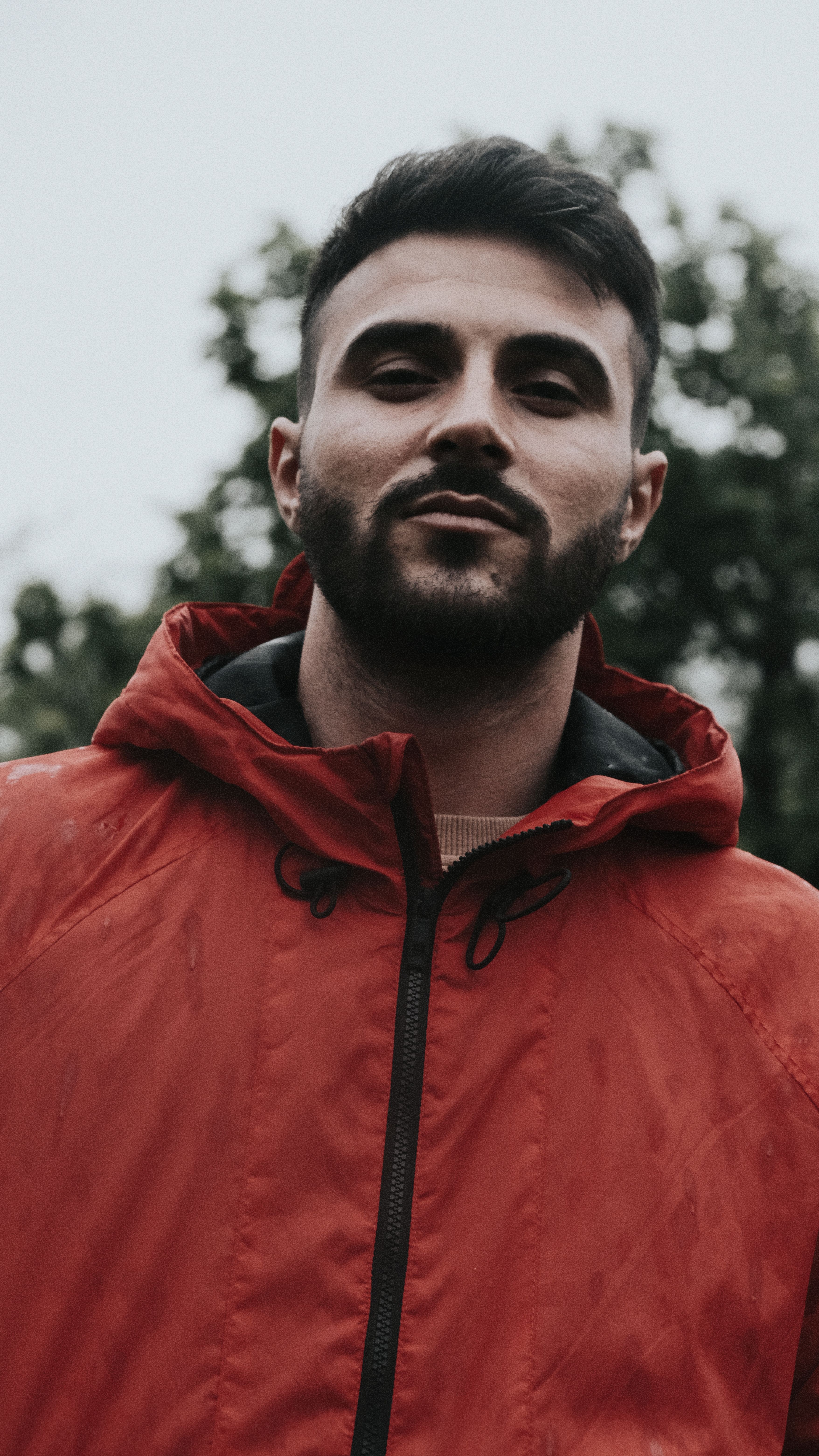 A confident man with a beard in a red jacket standing outdoors. Moody atmosphere captured with natural lighting.