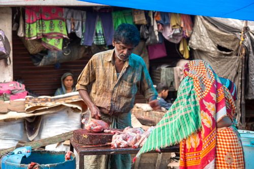 Free A man skillfully cuts meat at a bustling outdoor market filled with colorful fabrics and busy shoppers. Stock Photo
