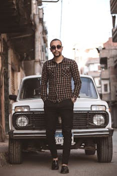 A fashionable man in sunglasses stands confidently against a vintage car in a city alley.