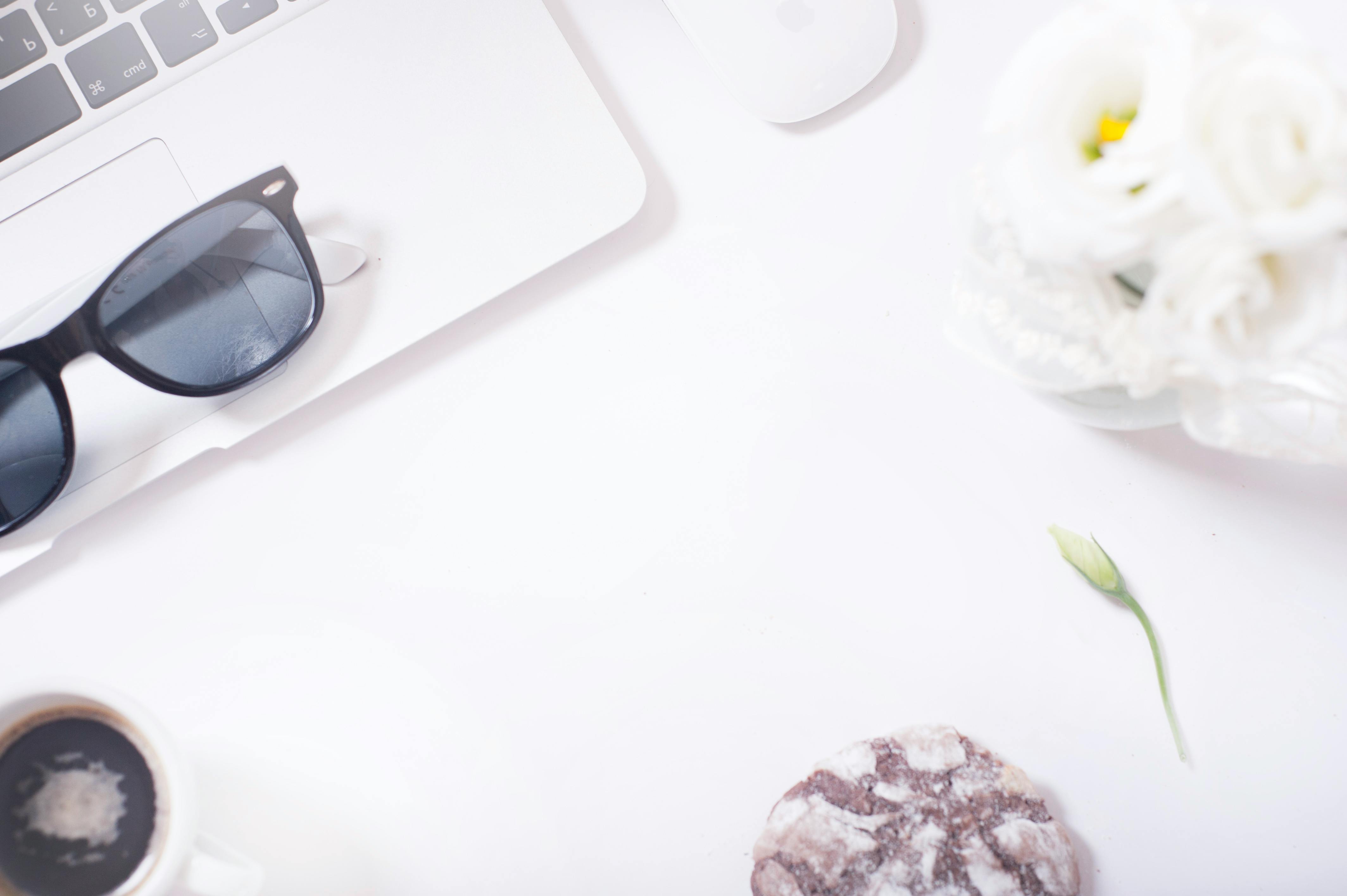 A minimalist and stylish workspace featuring sunglasses, flowers, and a laptop creating a bright aesthetic.