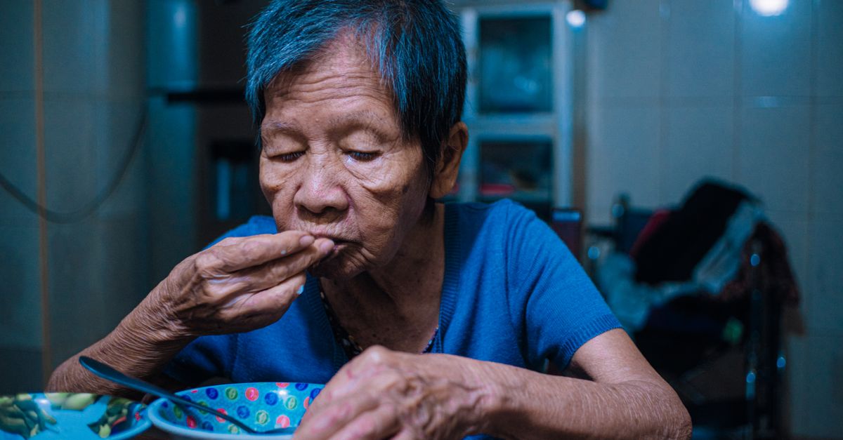Elderly Asian woman in a blue top enjoys a meal indoors, showcasing cultural dining practices.