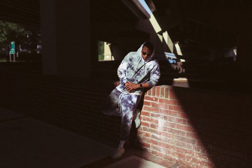 Free Man in tie-dye hoodie and pants leaning against a brick wall, under sunlight. Stock Photo