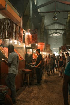 A lively night bazaar scene with people exploring vibrant street food stalls indoors.