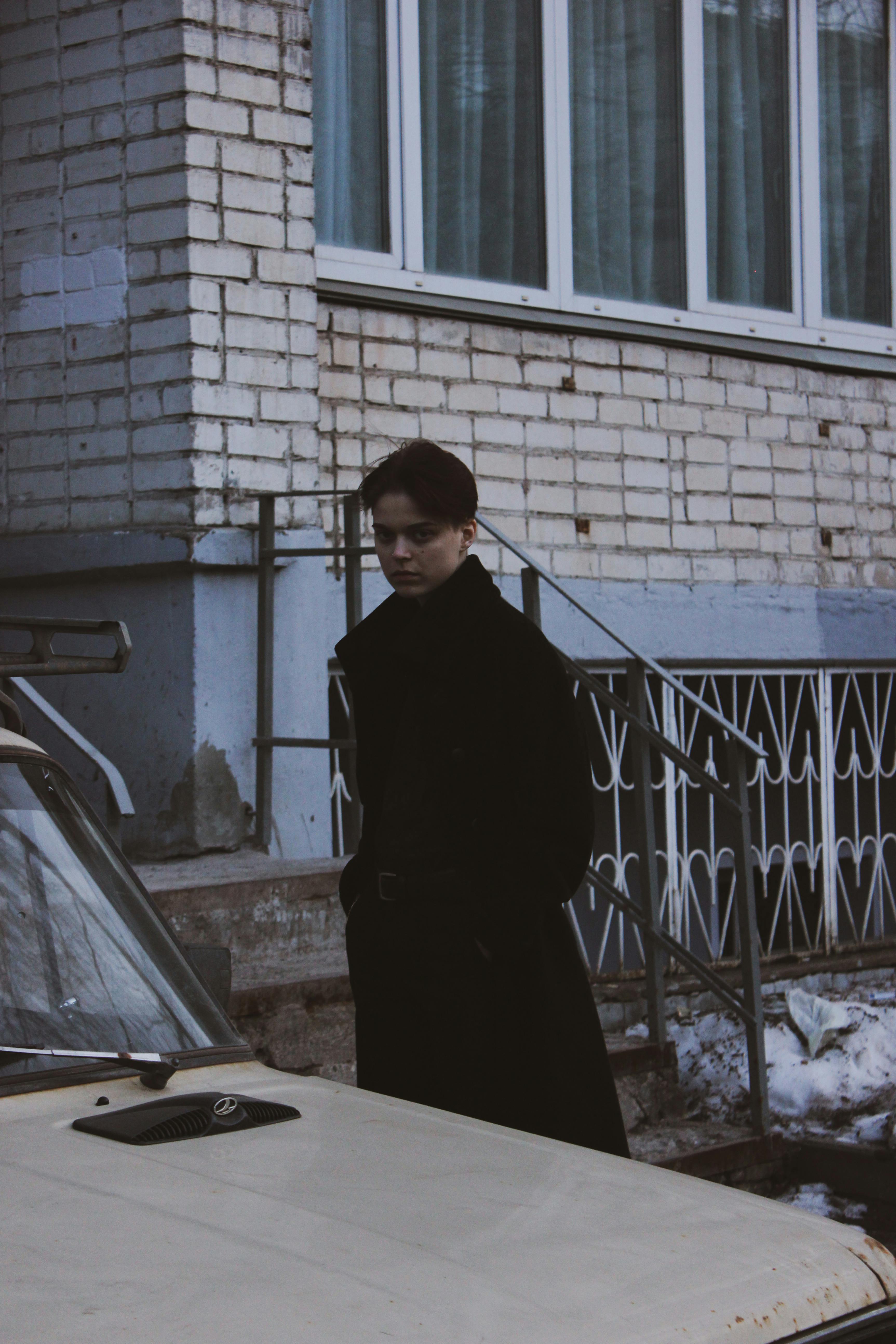 A man in a black coat stands by an old white car, exuding a moody vibe.
