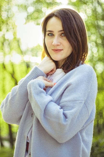 A woman posing outdoors in a soft blue cardigan with a serene expression, surrounded by greenery.