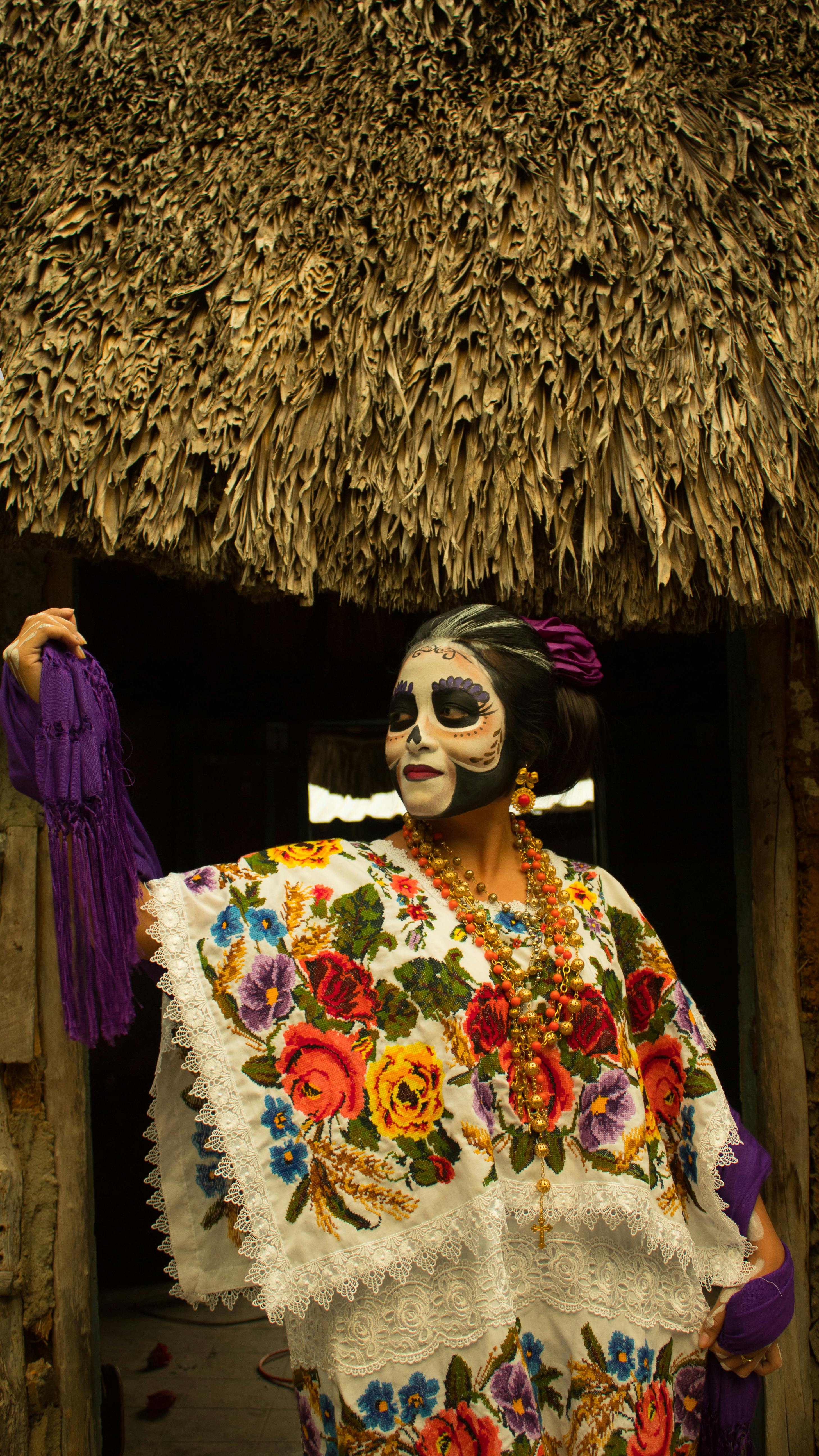 A woman in vibrant traditional Dia de Muertos attire poses in Yucatán, Mexico, showcasing colorful floral embroidery and makeup.