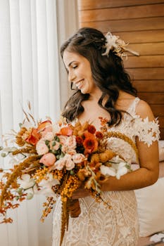 Bride in lace dress holding a stunning autumn-colored floral bouquet, smiling indoors.
